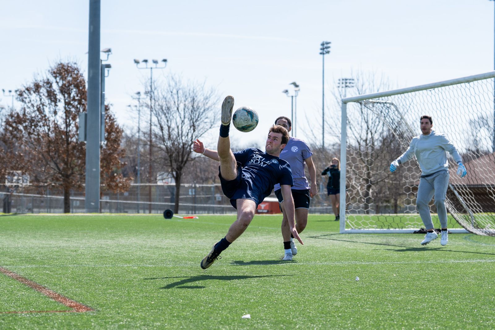 A player in navy strikes an overhead kick as the goalkeeper watches